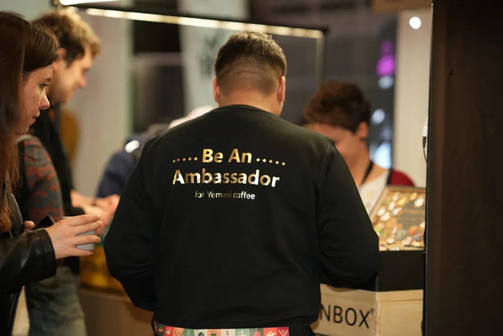 Person wearing a t-shirt that says "Be an Ambassador for Yemeni Coffee" at a coffee festival in Amsterdam.