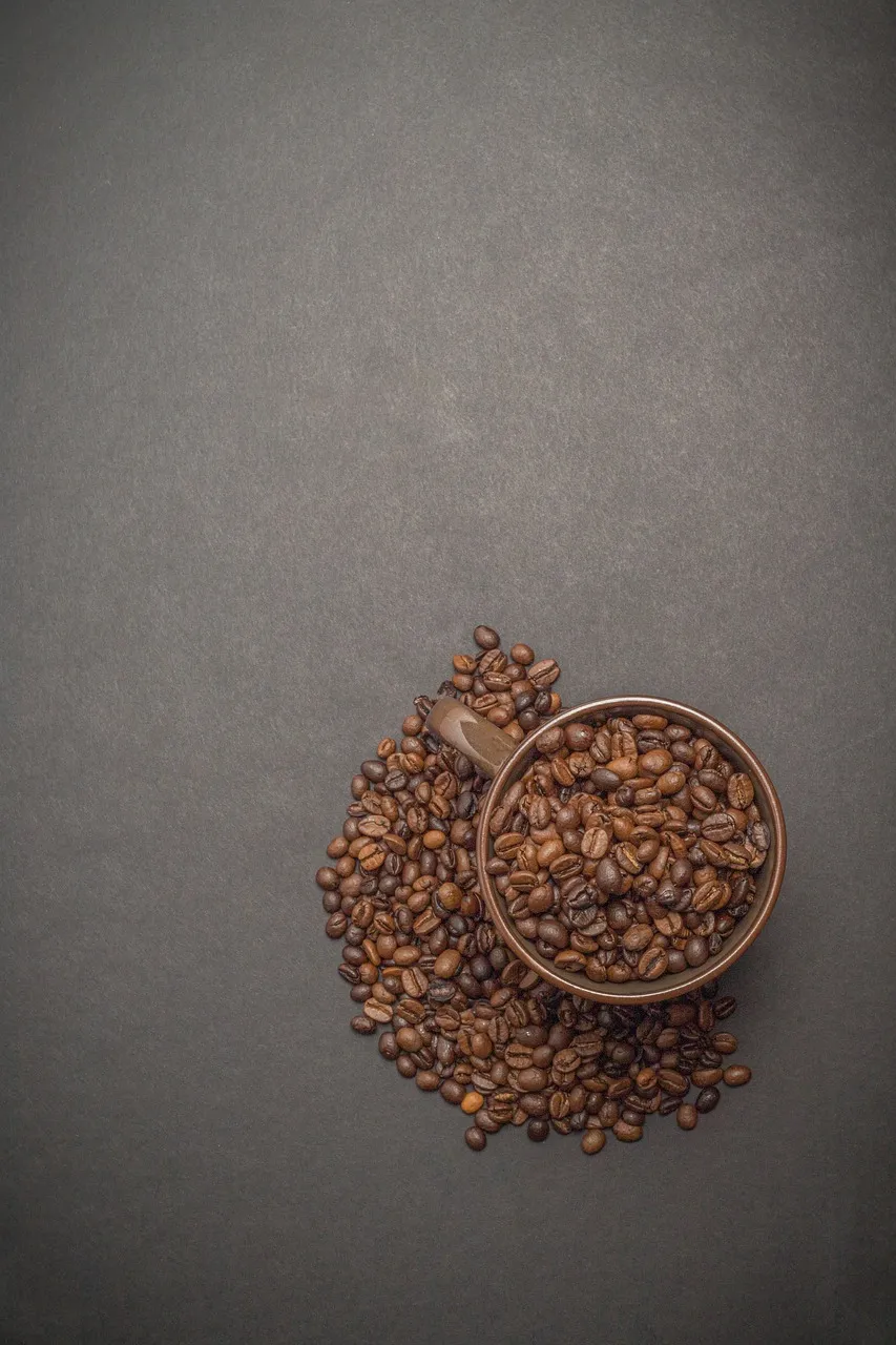 Top view of a brown cup filled with roasted coffee beans, with additional beans spilled around the cup on a dark surface.