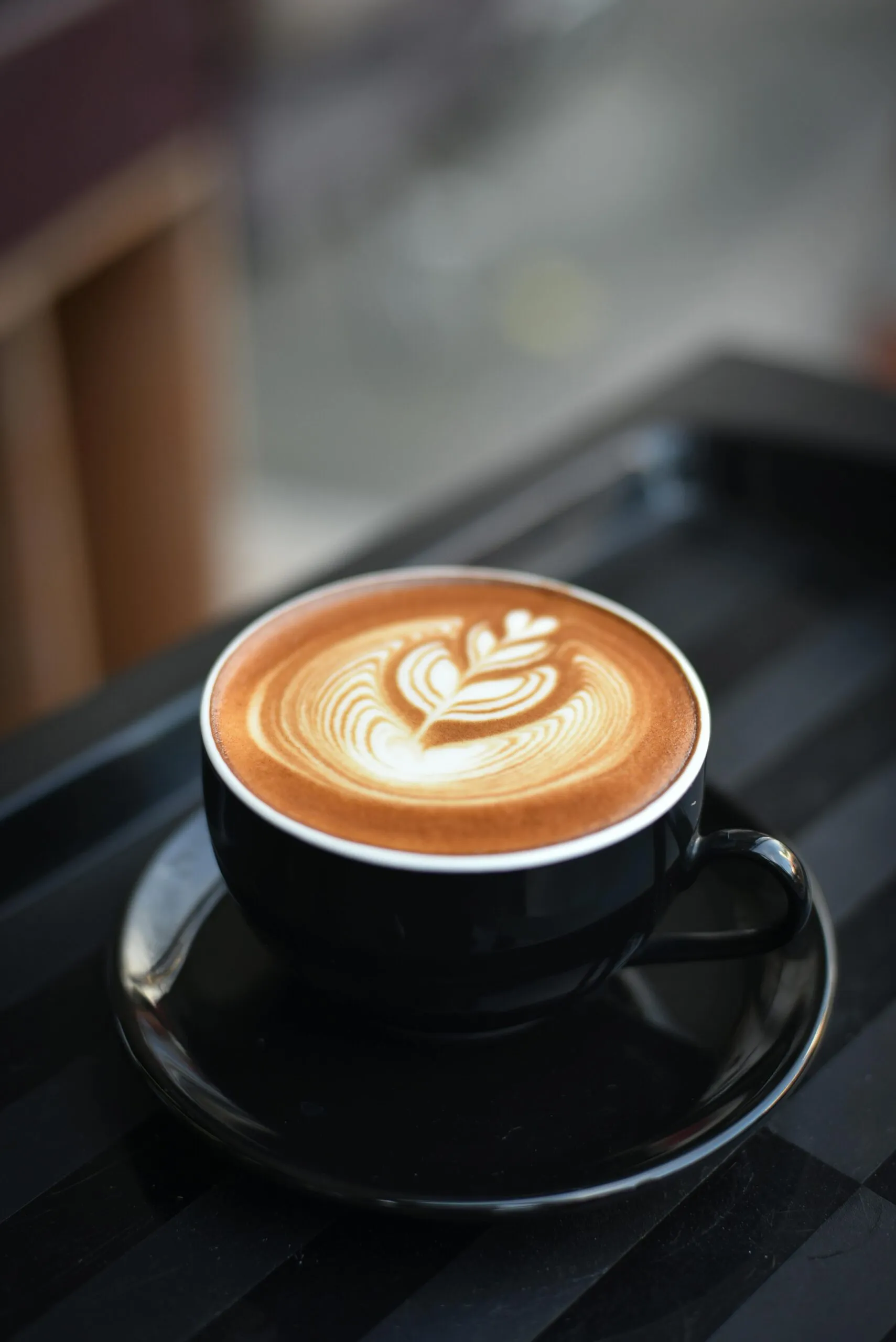 Latte art in a black cup with intricate barista design, set against a blurred background of a coffee shop interior.